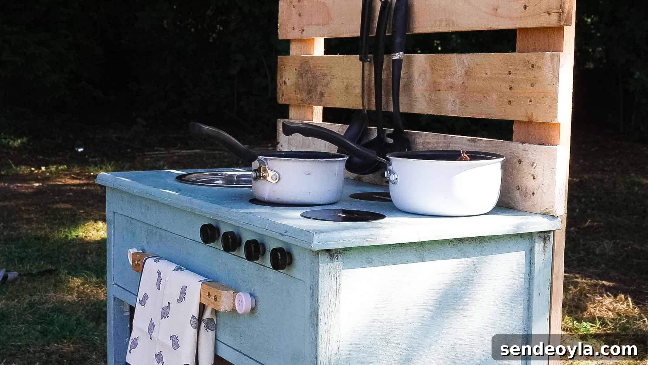 A pretend and rustic DIY small outdoor kitchen with pallet utensil holder with old pans on CD hobs, washing up bowl, drawer, knobs and tea towel