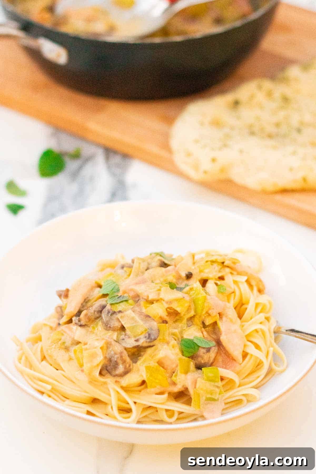 Cheesy Leek and Bacon Linguine 7 Close up of creamy cheese and bacon pasta in a large white bowl with pan full of leftovers on a chopping board and garlic bread